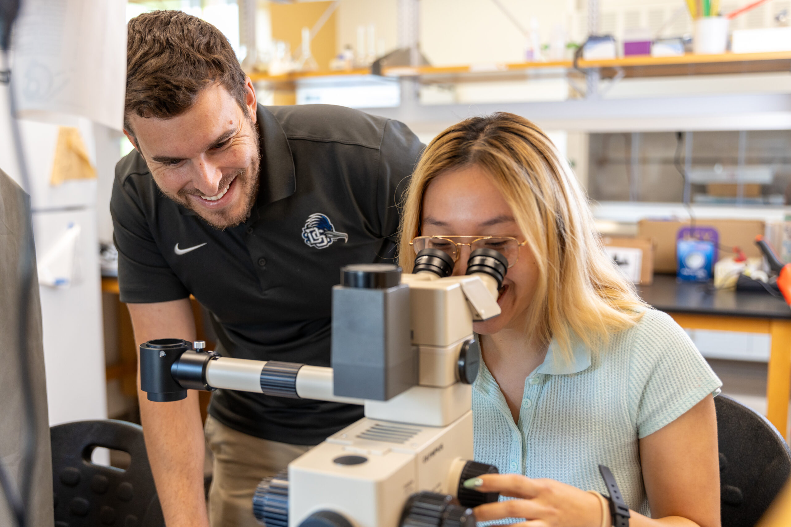 Female Dalton state student looks through microscope while male Dalton state teacher smiles while watching her.